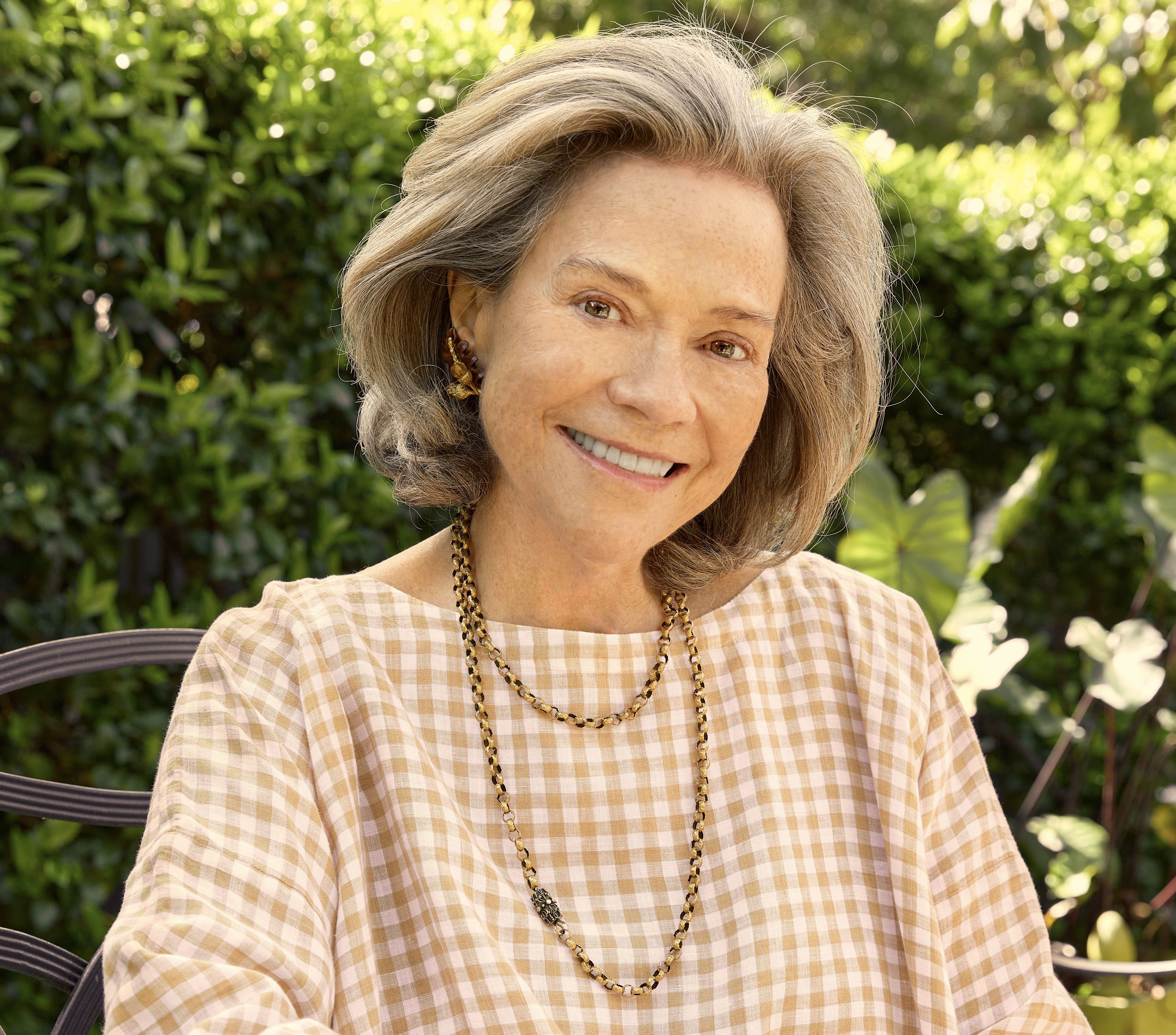 Headshot of Suzanne Rheinstein, seated in a garden wearing a gingham top.